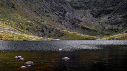 Coomloughra Lough reflects the surrounding rugged mountains. Soft ripples disrupt the peaceful surface, Irish landscape.