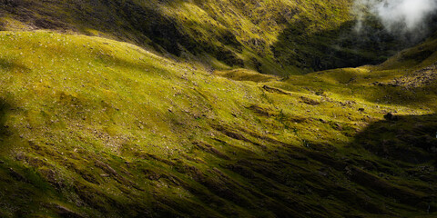 Sunlight illuminates grassy slopes in the mountainous. Patches of shadow are visible and low-lying clouds can be seen near the mountain peaks.