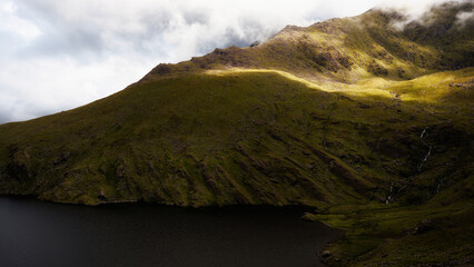 A scenic vista captures the dark waters of Lough Callee nestled against the lush, green slopes of mountains. Clouds drape over the peaks and sunlight breaks through.