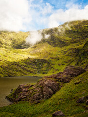 Green hillsides lead down to Lough Callee. Mist hangs on the upper reaches of the hills, with blue sky visible above. Rocks are visible along the lakeside.
