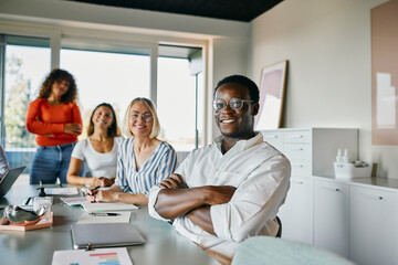 Diverse Team Enjoying a Productive Meeting in Modern Office Environment. They are all looking directly into the camera while smiling. On the table are laptops and notebooks.