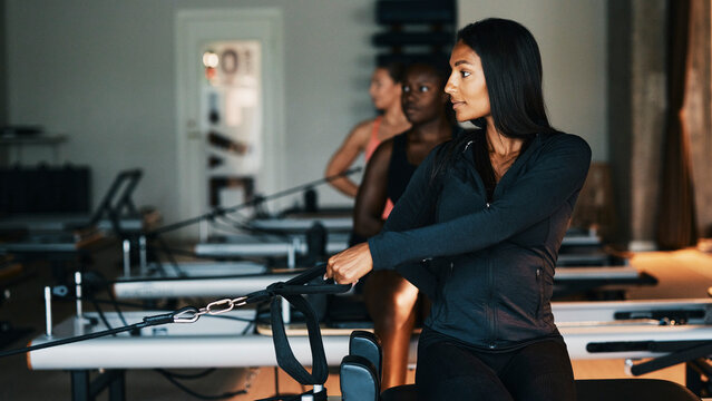 Fit young diverse woman in sportswear using resistance bands on a reformer during a workout class with other women at a pilates gym