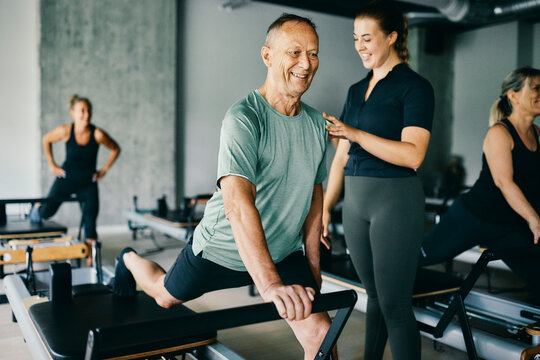 Smiling fitness instructor working with a mature male client during a reformer class with a group of people in a pilates gym
