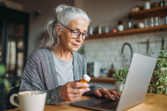 Senior woman using laptop computer to research medication refills online from home kitchen - Powered by Adobe