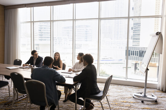 Busy multicultural team investors executives wear formal suits discuss work at modern city view panoramic windows boardroom. Diverse stakeholders group hold negotiations together in round table format