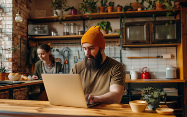  Focused man in orange beanie working on laptop at rustic kitchen. Cozy freelance work concept.  
