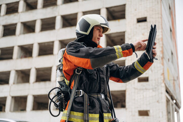 Fototapeta premium Tablet in hands. Professional firefighter is standing outdoors against big unfinished building