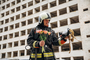 Beautiful flowers in hands. Professional firefighter is standing outdoors against big unfinished...