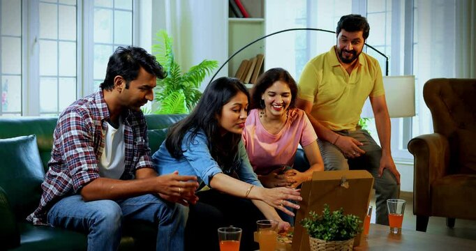 Friends having pizza at home, Indian Asian young close friends sitting together on sofa in cozy modern house, smiling and enjoying cheesy slices while showing pizza box or slice to camera with joy