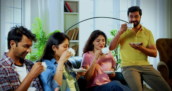 Friends enjoying tea at home, Indian Asian young close friends drinking hot chai or coffee while relaxing on sofa in cozy modern home, smiling and holding cups up to camera to show warm hospitality