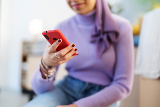 A young woman wearing a purple hijab is using a red smartphone. She is looking at the screen and interacting with the device. This image shows modern technology use and connectivity.