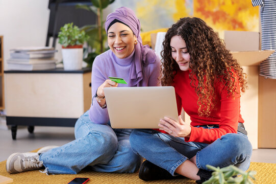 Two young women sit on the floor using a laptop and credit card for online shopping or banking. They are smiling and enjoying the process together.