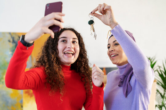 Two diverse young women celebrate a significant milestone, holding up keys and capturing the moment with a smartphone selfie indoors. They are happy.