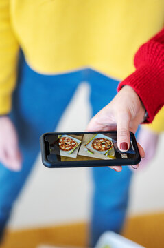 Close up of a hand holding a smartphone. The phone screen displays photos of two delicious pizzas. A person in a red and yellow outfit is capturing the food image.