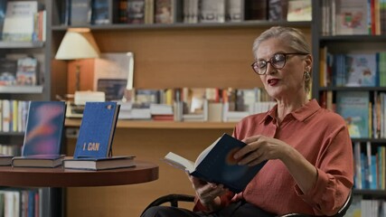 Medium slowmo of mature female author looking at camera as reading aloud from her new book in intimate bookstore setting during literary event or signing session