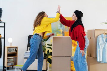 Two diverse women high-five, celebrating success or a milestone like moving into a new home. Cardboard boxes are visible in the background.