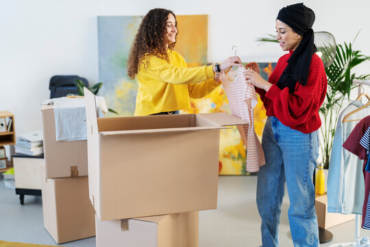 Two diverse women pack clothes into cardboard boxes. They are sorting through garments, preparing for a move or donation. This shows friendship and teamwork.