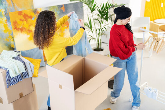 Two women are packing clothes into cardboard boxes. One woman puts a shirt in a box while the other organizes hangers on a rack. They are moving or decluttering their home.