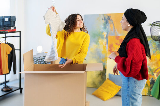 Two friends are packing clothes into a large cardboard box. One friend holds up a striped shirt from the box while the other looks on. They are smiling and sharing a moment.