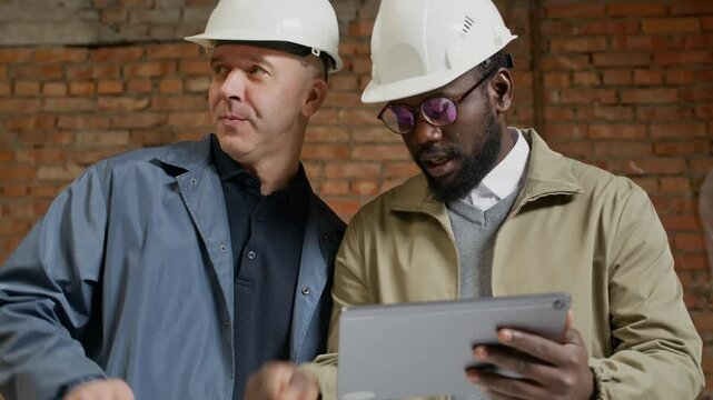 Tilt up shot of two male bicultural engineers in hardhats using tablet PC while discussing renovation plan on construction site