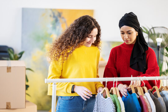 Two diverse young women are browsing clothes on a rack in a bright room. They are choosing items from the rail, possibly for sale or personal use.