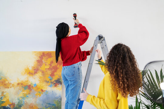 Two women work together on home improvement. One woman stands on a ladder using a power drill to make a hole in the wall while the other assists her. They are preparing to hang artwork.