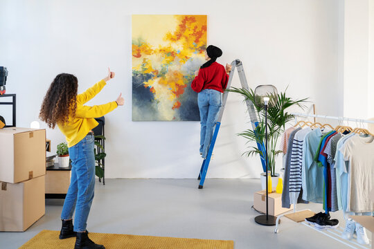 Two young women decorate their new home. One hangs a large abstract painting on the wall from a ladder while the other gives a thumbs-up gesture, approving the placement.