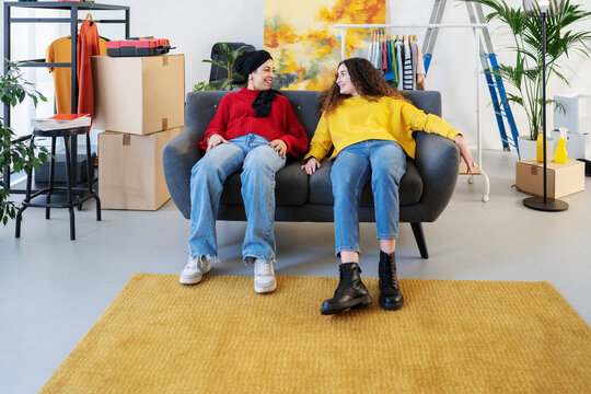 Two young women, one in a hijab, sit on a sofa in a room filled with moving boxes and items. They smile, taking a break from setting up their new space.