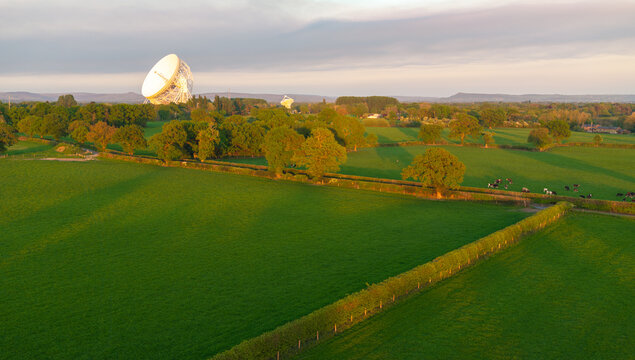 Panoramic aerial image of green landscape of British countryside with a white radio telescope dish and Cows grazing on a fields.  - Powered by Adobe