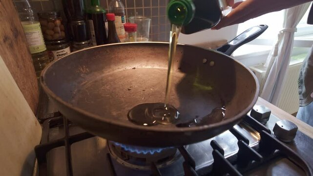 Domestic chef pouring olive oil into hot frying pan on kitchen gas hob