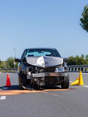 A damaged car sits on the highway under a clear blue sky surrounded by traffic cones. Emergency services are on the way to manage the situation and clear the road.