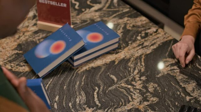 Closeup of bookstore counter as employee handing over blue book to customer holding credit card after making payment, with stack of books and red bestseller table tent in background