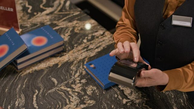 Close up of transaction at bookstore counter as customer paying with card and employee holding payment terminal and handing blue, hardcovered purchase beside stacked books