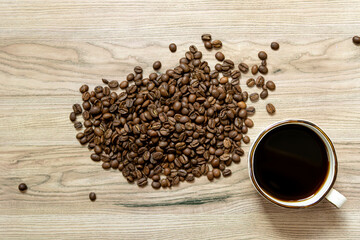 A cup of black coffee and scattered roasted coffee beans on a light wooden surface. The coffee beans are placed in the center and to the left, while the cup is on the right. 