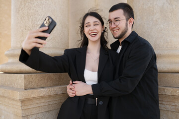 Happy young couple taking a selfie near stone columns. The woman laughs while the man hugs her from behind. Candid joyful moment of modern love in an urban setting.