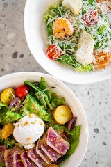 Fresh gourmet salads in bowls. One features shrimp, cherry tomatoes, croutons, and grated cheese on lettuce. The other includes seared tuna, a poached egg, cherry tomatoes, olives, roasted potatoes