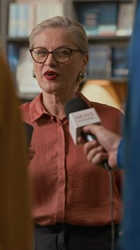 Vertical over-the-shoulder shot of mature Caucasian female author speaking into news-branded microphone during interview at bookstore event, addressing media following her autograph session