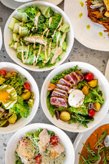 A selection of fresh gourmet salads in white bowls on a gray countertop, featuring ingredients like seared tuna, poached eggs, cherry tomatoes, mixed greens, and vibrant vegetables.