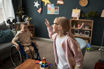 Two children, one in wheelchair, interacting and smiling in cozy living room setting with toys and decorations visible. Both kids joyfully engaged, creating warm atmosphere