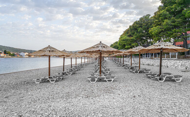 A serene pebble beach lined with rustic straw parasols under a blue sky beside calm sea waters.