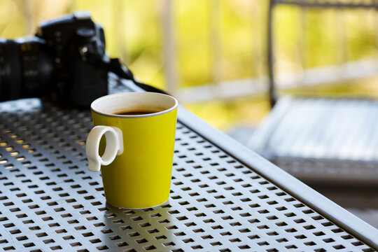 A yellow mug of coffee on a metal outdoor table with blurred camera and chair in background