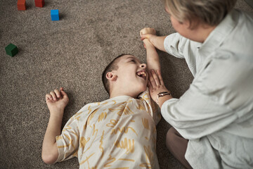 Child with disability lying on carpet with arm extended, holding caregiver's hand during exercises. Interaction appears playful and heartwarming