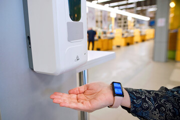 Closeup of female hand activating touchless hand sanitizer dispenser in shopping center, demonstrating modern hygiene practices and public safety precautions. Wireless automatic antiseptic dispenser