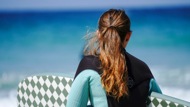 A woman wearing a wetsuit holding a patterned surfboard looks out towards the ocean. The blue water and clear sky create a serene and adventurous seaside atmosphere at this popular Cornish resort.