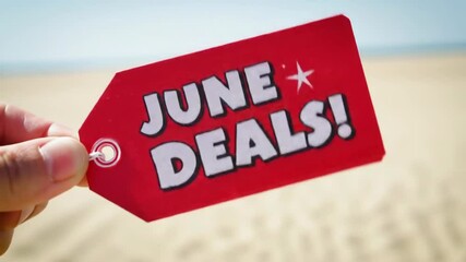 Hand holding "JUNE DEALS!" promotional tag against sandy beach background  