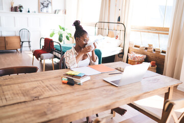 Woman working remotely from stylish home interior, reaching toward laptop with one hand while sitting at rustic desk filled with planners, markers, phone and digital accessories