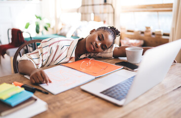 Close-up of peaceful woman napping on wooden table with notebook and laptop, glasses resting on open page, enjoying quiet break in warm, homey environment