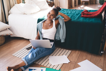 Relaxed young black woman sitting on floor, laptop on legs, leaning on bed and daydreaming with soft smile during break from remote freelance work at home