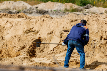 A man in a blue jacket and jeans diligently works with a shovel, digging sand at a construction site.