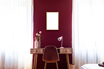 Elegant wooden desk with velvet chair, lamp, and empty frame on burgundy wall. Ideal for mockups and personalization.